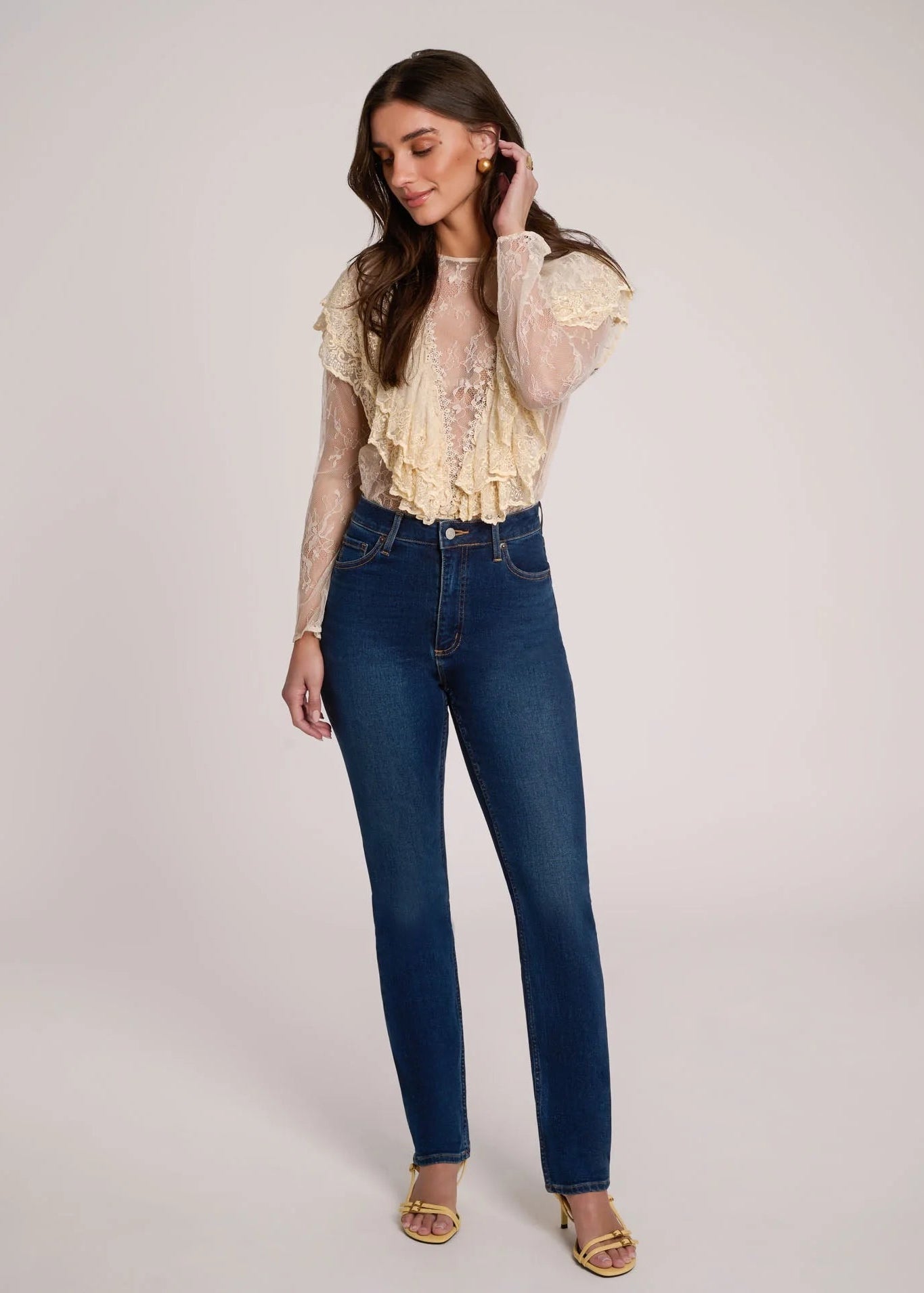 A woman modeling a cream ruffled lace long-sleeve top paired with high-waisted dark blue skinny jeans and gold sandals, standing against a light gray studio background.