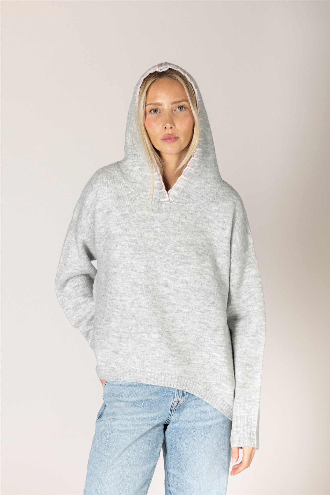 A woman wearing a penny pullover from Lyla and Luxe gray heathered knit pullover hoodie with a wide collar and drawstring hood, paired with light blue jeans, against a plain light gray studio background.