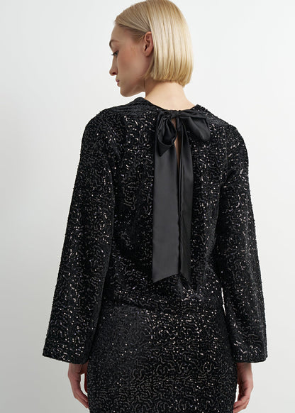 A woman shown from the back wearing a black long-sleeve sequined dress with a large satin bow at the neckline, against a plain light gray studio background.