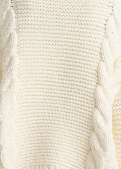 Close-up of a cream-colored cable-knit sweater showing the detailed knit texture and twisted cable patterns.