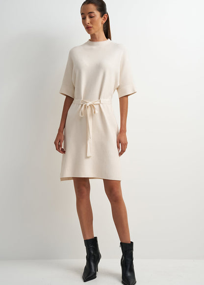 A woman modeling a cream-colored short-sleeve knit dress with a matching tie belt at the waist, paired with black ankle boots, against a plain light gray studio background.
