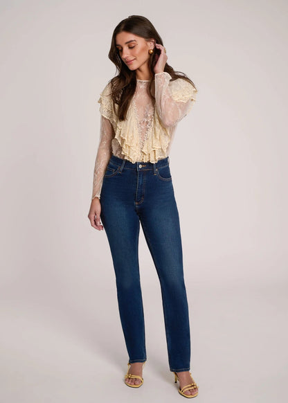A woman modeling a cream ruffled lace long-sleeve top paired with high-waisted dark blue skinny jeans and gold sandals, standing against a light gray studio background.