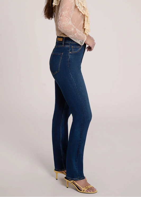 A woman stands in profile wearing a cream sheer lace ruffle blouse, dark blue high-waisted flare jeans, and gold heeled sandals against a plain light gray studio background.
