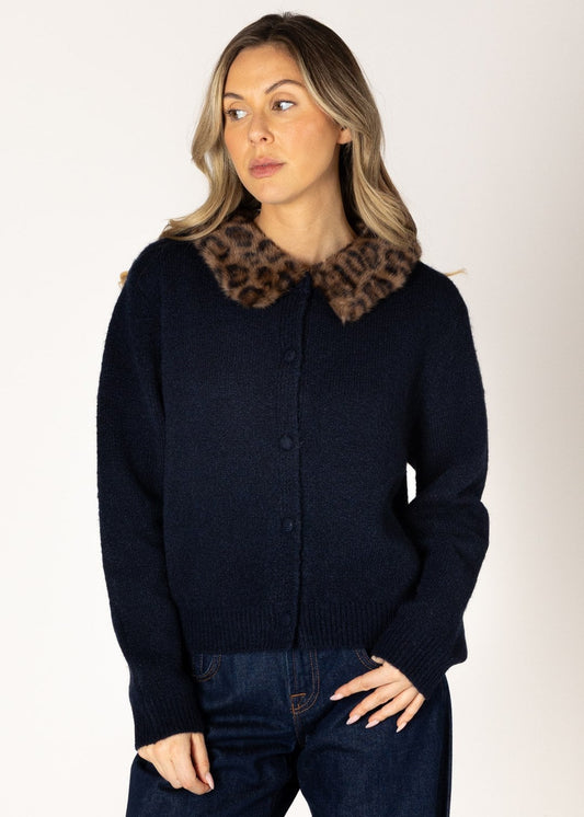 A woman modeling a navy blue knit cardigan featuring a large brown and black leopard-print collar, paired with dark blue jeans, against a plain light gray studio background.