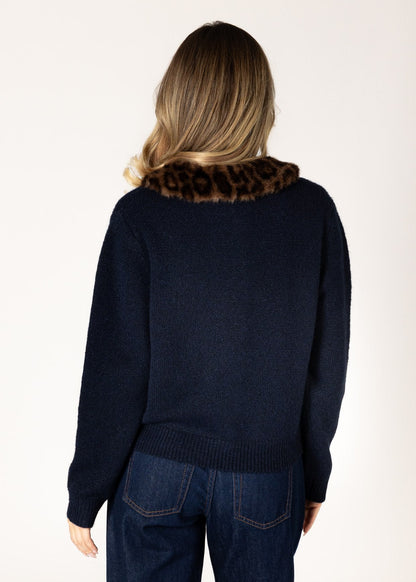 A woman viewed from behind wearing a navy blue knit sweater with a brown and black leopard-print faux fur collar, paired with dark blue jeans, against a plain off-white background.