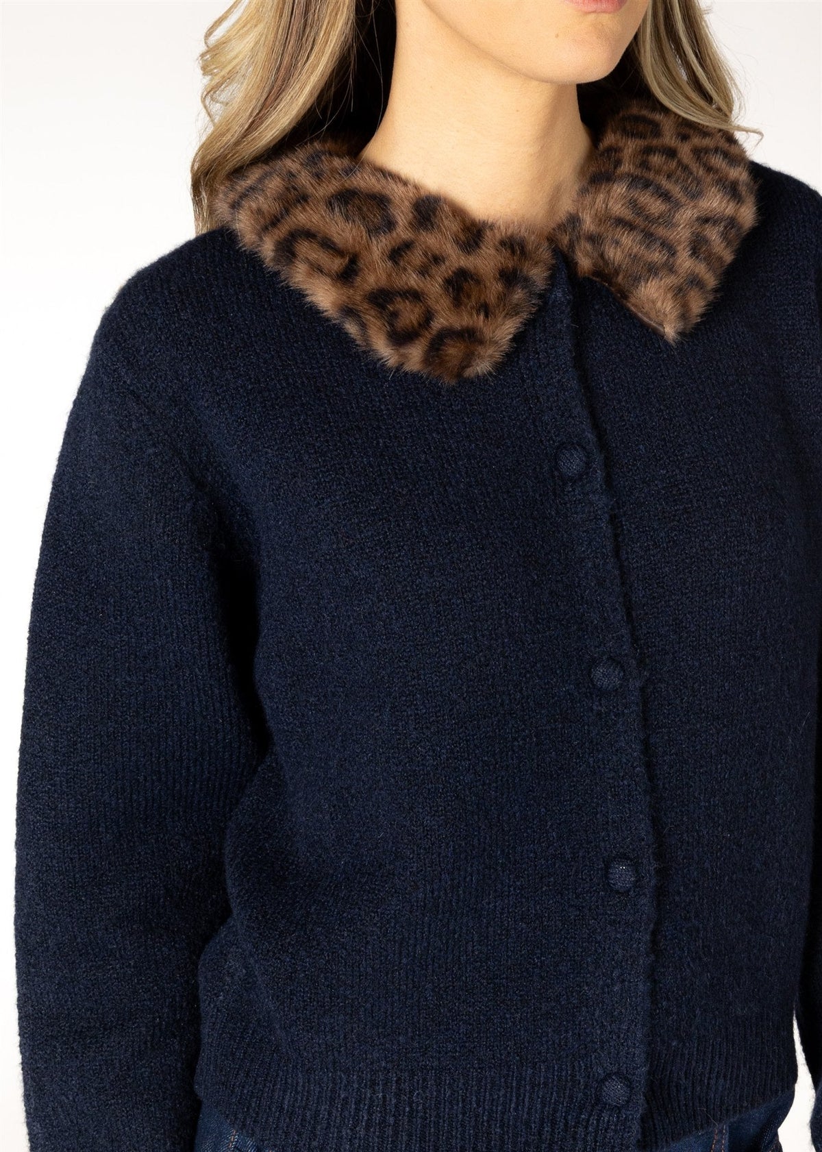 A close-up of a person wearing a dark navy knit cardigan with a large brown and black leopard-print faux fur collar, paired with blue jeans against a plain white background.