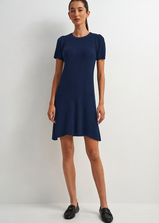 A woman modeling a short-sleeve, crew-neck ribbed knit dress in navy blue, paired with black loafers, against a plain light gray studio background.