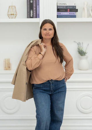A woman stands in a bright white room with built-in bookshelves, wearing a beige long-sleeve top, blue jeans, and carrying a tan coat over her shoulder.