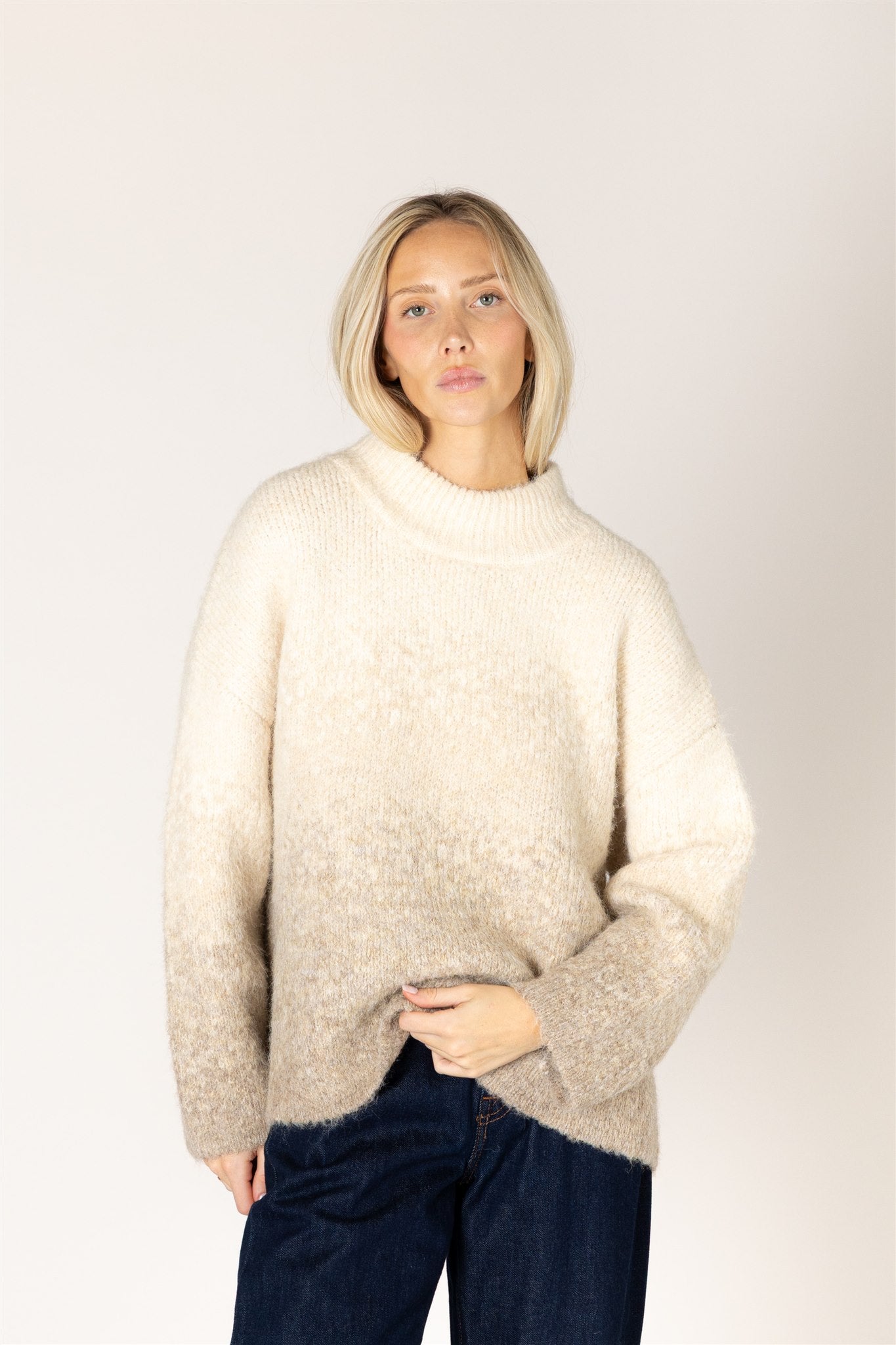 A woman with blonde hair wearing a fuzzy, oversized cream-colored knit sweater and dark blue jeans, standing against a plain light gray studio background.