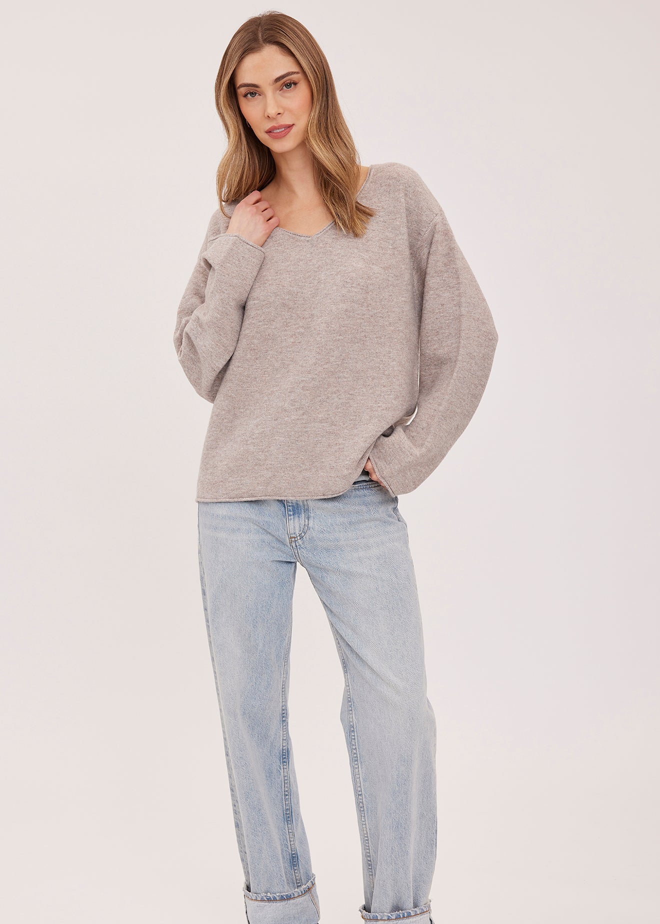 A woman modeling a light heather gray oversized knit sweater, paired with light-wash blue jeans and black slip-on shoes, against a plain light gray studio background.