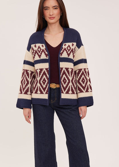 A woman modeling a navy blue knit cardigan with cream and burgundy geometric patterns, paired with dark blue wide-leg jeans, a burgundy top, and beige ankle boots against a light beige studio background.