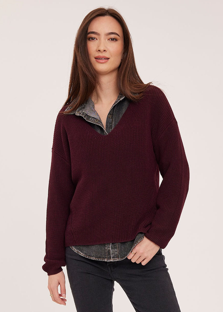 A woman modeling a dark burgundy knit sweater with a layered gray shirt underneath, paired with dark gray jeans and brown ankle boots, against a light gray studio background.