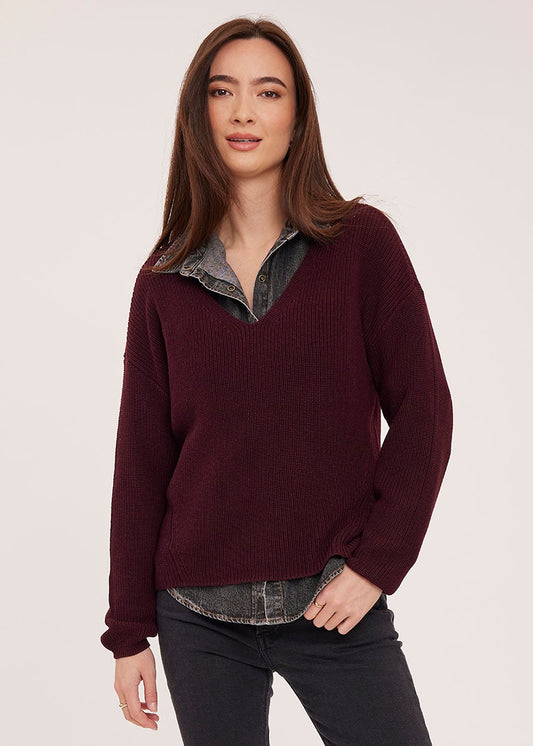 A woman modeling a dark burgundy knit sweater with a layered gray shirt underneath, paired with dark gray jeans and brown ankle boots, against a light gray studio background.