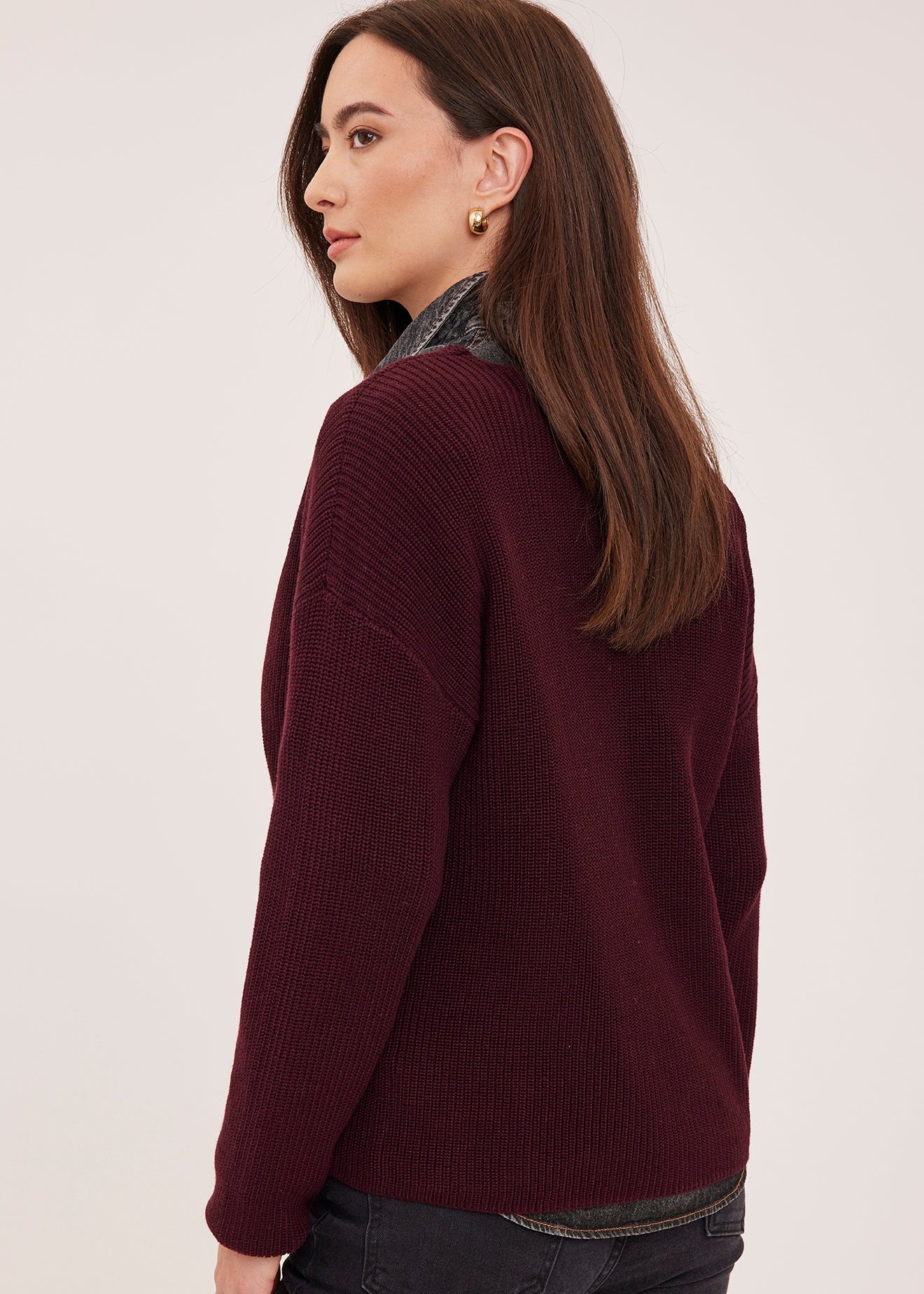 A woman shown from the back-three-quarter view wearing a ribbed knit sweater in deep burgundy over a dark patterned shirt and dark jeans, against a plain light background.