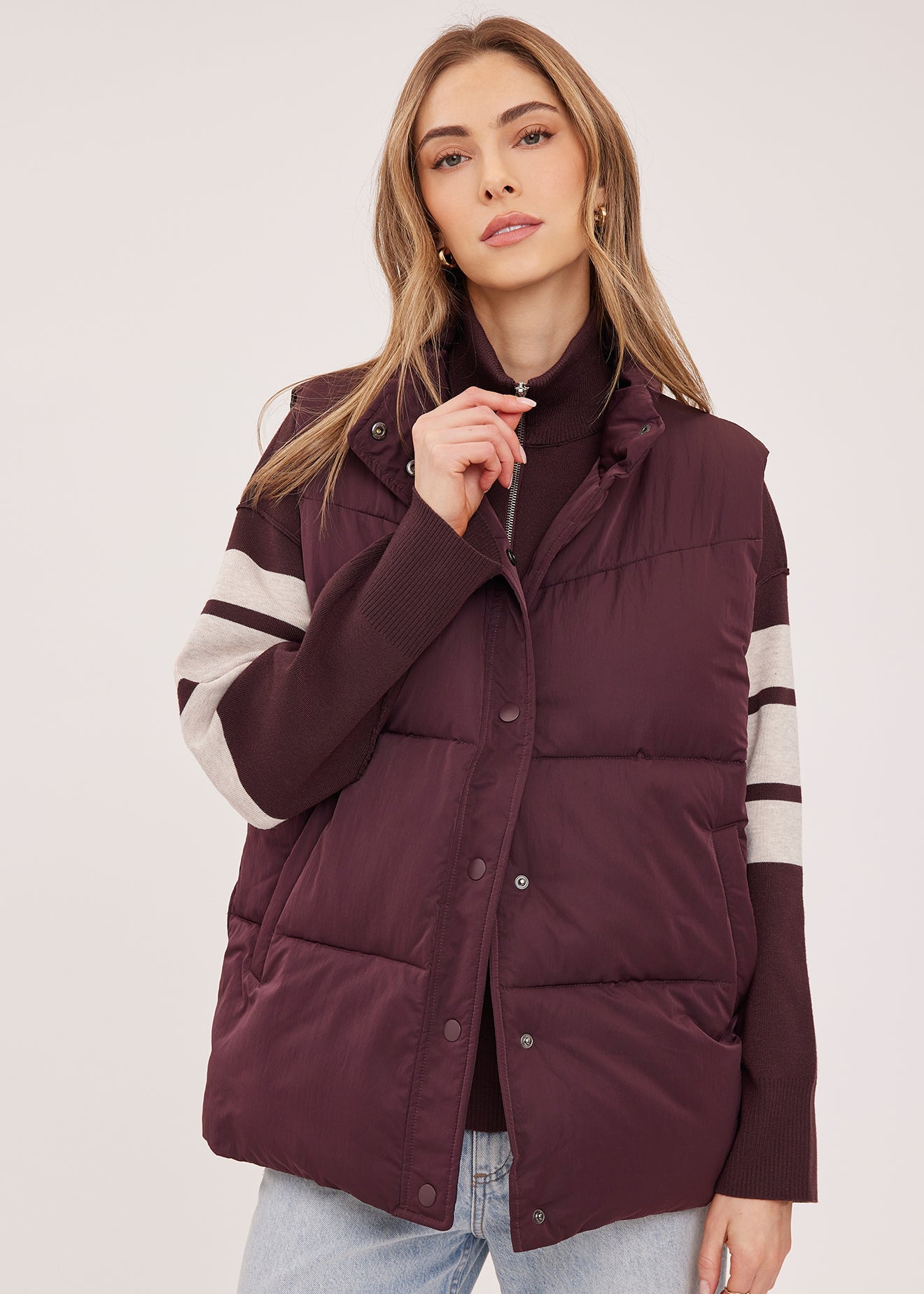 A woman wearing a sleeveless puffer vest in deep burgundy with cream stripes on the sleeves, styled over a black turtleneck and light wash jeans against a plain light background.