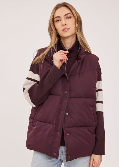 A woman wearing a sleeveless puffer vest in deep burgundy with cream stripes on the sleeves, styled over a black turtleneck and light wash jeans against a plain light background.