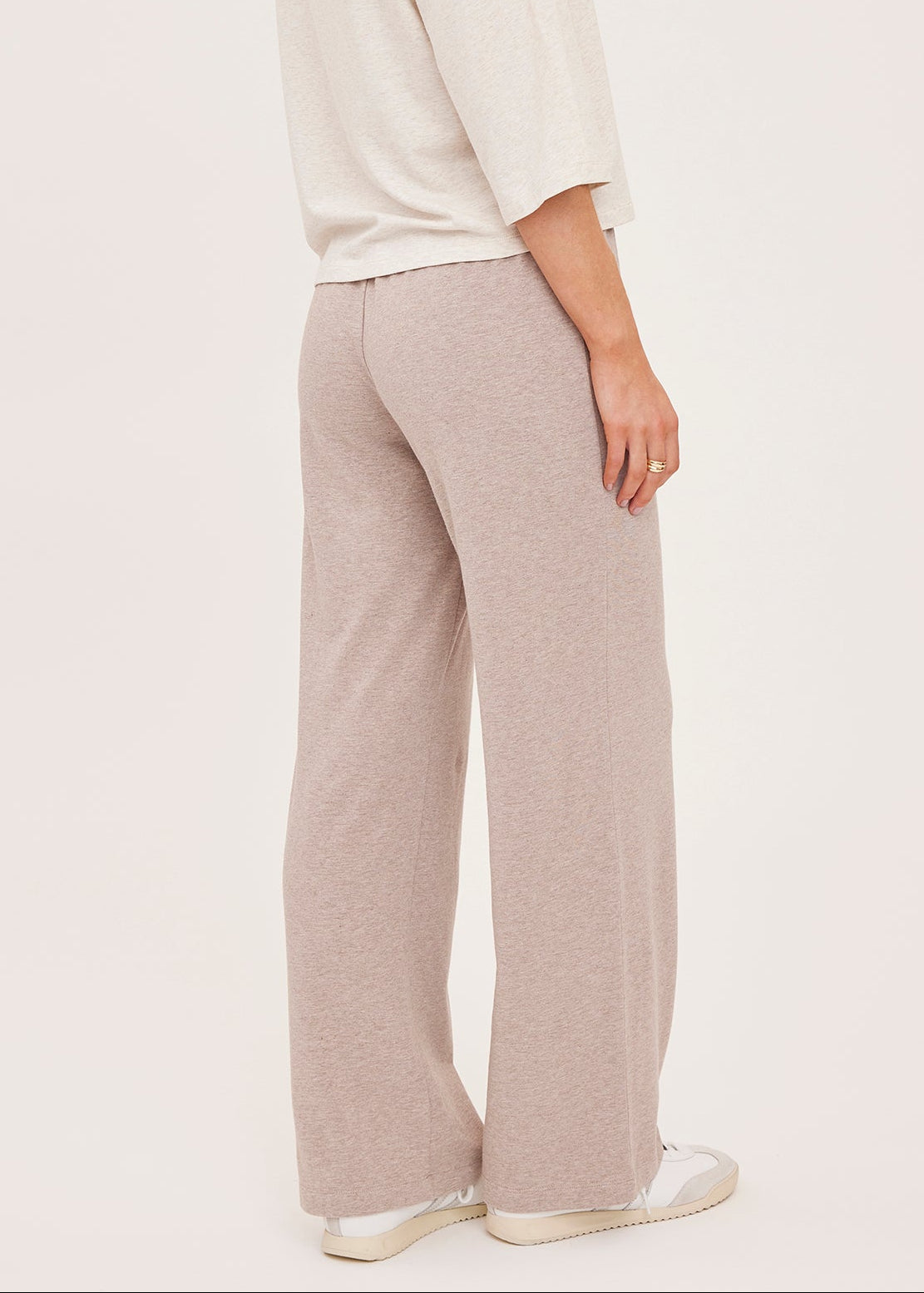 A woman shown from the back wearing a cream three-quarter sleeve top and light beige wide-leg knit pants, paired with white sneakers, against a solid off-white background.