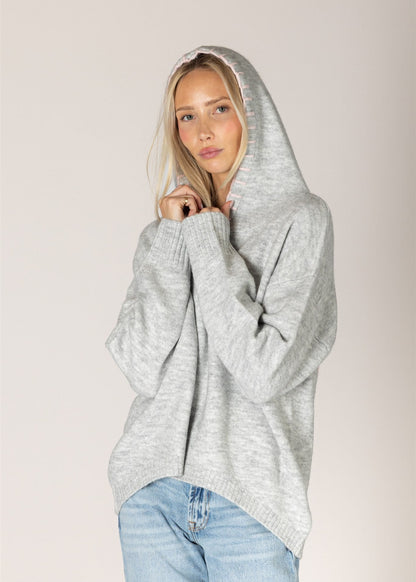 A woman wearing a light gray heathered oversized knit hoodie sweater and light blue jeans, posing against a solid light beige studio background.