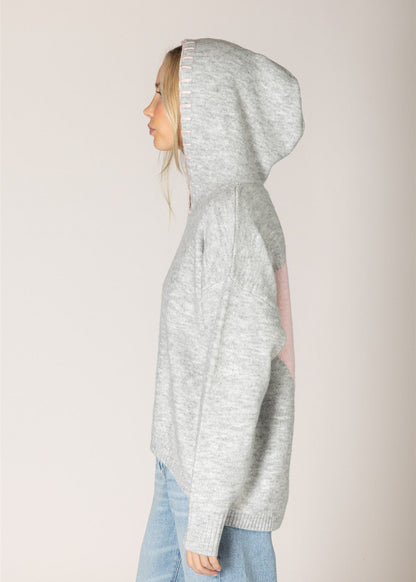 A woman shown in profile wearing a light heather gray knit hoodie with a pink patch on the back, paired with light blue jeans, against a solid light beige background.