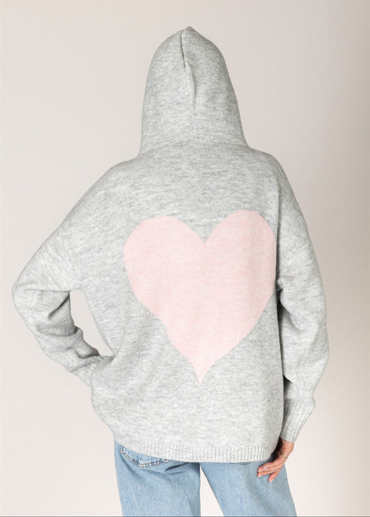 A person shown from the back wearing a light gray heathered hoodie with a large light pink heart patch on the back, paired with light blue jeans, against a plain light beige background.