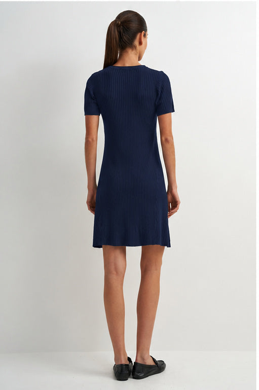 A woman viewed from the back wearing a short-sleeve ribbed knit dress in dark navy blue, paired with black flats, against a plain light gray studio background.
